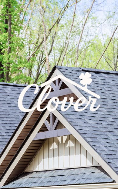 A house roof with gray shingles, exposed beams, and trees in the background. The words "Clover Roofing" and a clover icon are overlaid at the top, highlighting this trusted South Carolina roofing brand.