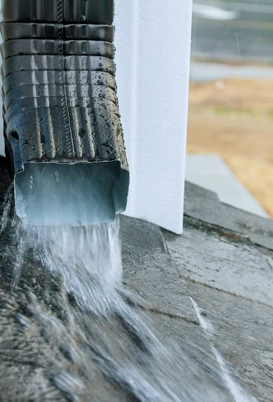 Water flows out of a metal downspout onto a wet stone surface near a building corner, showcasing the dependable workmanship of Clover Roofing in South Carolina.