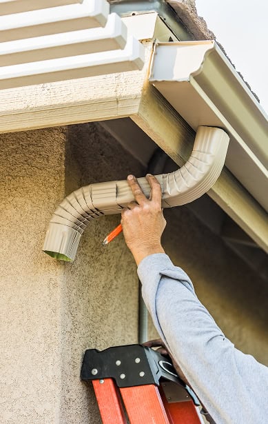 A person on a ladder installs or adjusts a downspout to the gutter system on the side of a house, holding a pencil and the downspout in place—expert service from Clover Roofing in South Carolina.