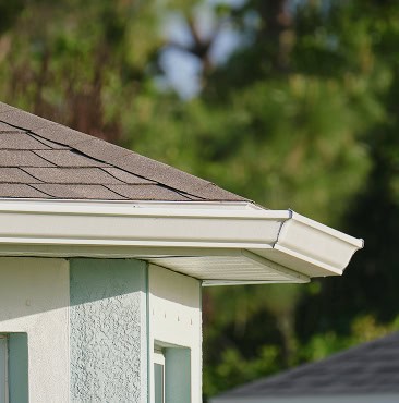 Close-up of a house corner with a white gutter attached to the roof edge, installed by Clover Roofing in South Carolina. Trees and another roof are softly blurred in the background.