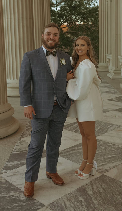 A man in a plaid suit and bow tie stands next to a woman in a white dress and heels on a marble walkway with columns in the background.