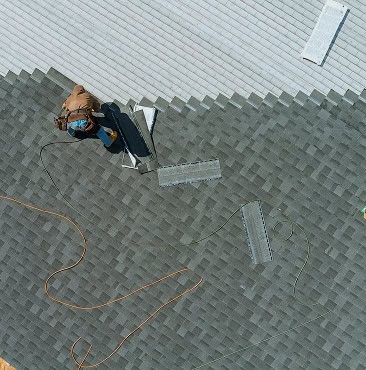 A person installs roofing shingles on a house roof, with tools, shingles, and extension cords visible nearby.