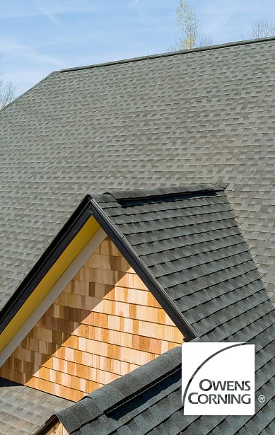 A house with a steep, dark gray asphalt shingle roof, natural wood siding, and the Owens Corning logo in the lower right corner—expertly crafted by Clover Roofing of South Carolina.