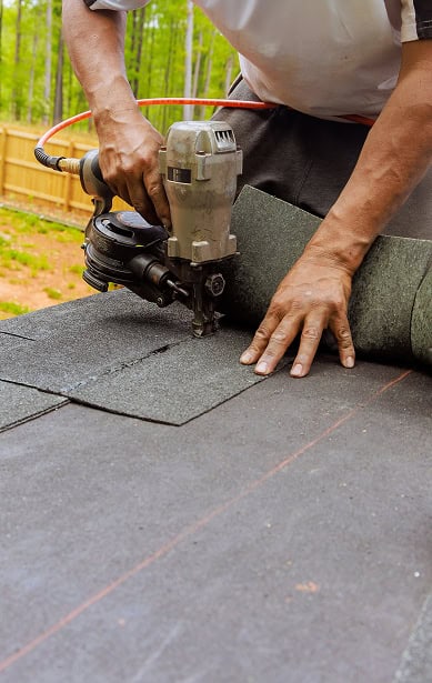 A person installs roofing shingles with a nail gun on a roof, with a roll of shingles and a wooden fence in the background, showcasing Clover Roofing expertise in South Carolina.