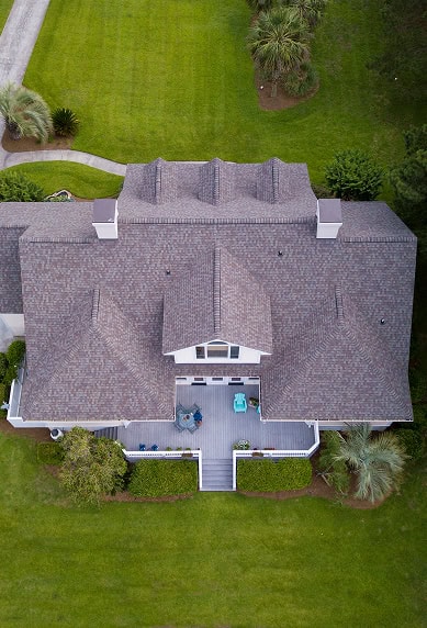 Aerial view of a two-story house with a large gray roof by Clover Roofing, featuring a front porch and surrounded by green lawn and trees in South Carolina.