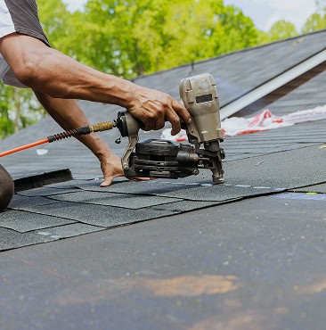 A person uses a nail gun to install asphalt shingles on a roof for Clover Roofing in South Carolina, with trees and blue sky visible in the background.