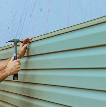 A worker from Clover Roofing installs light green vinyl siding on a house in South Carolina, using a hammer, with a layer of protective wrap visible above the siding panels.