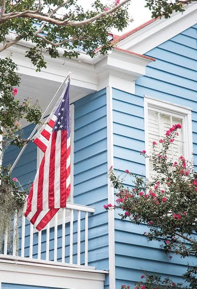 A blue wooden house with white trim, crafted by Clover Roofing, features an American flag hanging from the balcony. Pink flowers and green leaves frame part of this charming South Carolina scene.