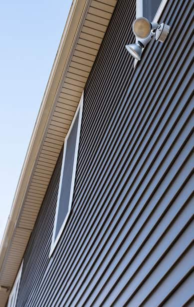 Close-up view of a house’s exterior with dark horizontal siding, a window, and a mounted outdoor floodlight under the roof’s eaves against a clear South Carolina sky—quality craftsmanship by Clover Roofing.