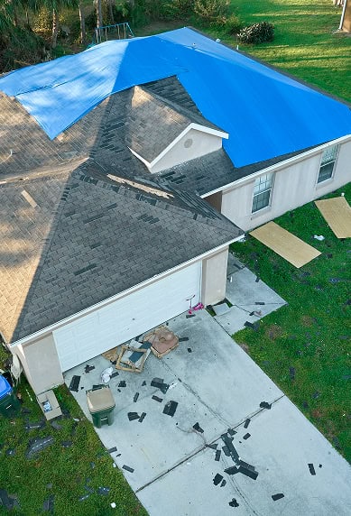 A house with a damaged roof partially covered by a blue tarp, with shingles scattered on the driveway and grass—a common sight for Clover Roofing after South Carolina storms.